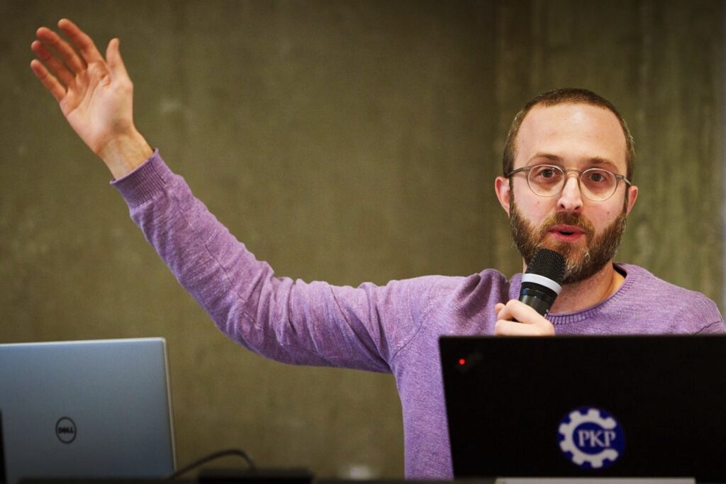 PKP's Nate Write, Software Developer, Workflow Lead, speaking passionately to an audience, with hand raised in the air and microphone in other hand. The laptop in front of Nate has a PKP sticker on it.