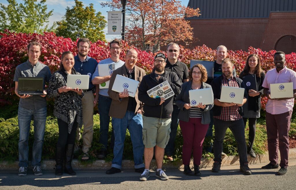 PKP's diverse, usually remote team, gathers together in a physical location outside for a group photo. All the members have PKP logos on their laptops in the photo.
