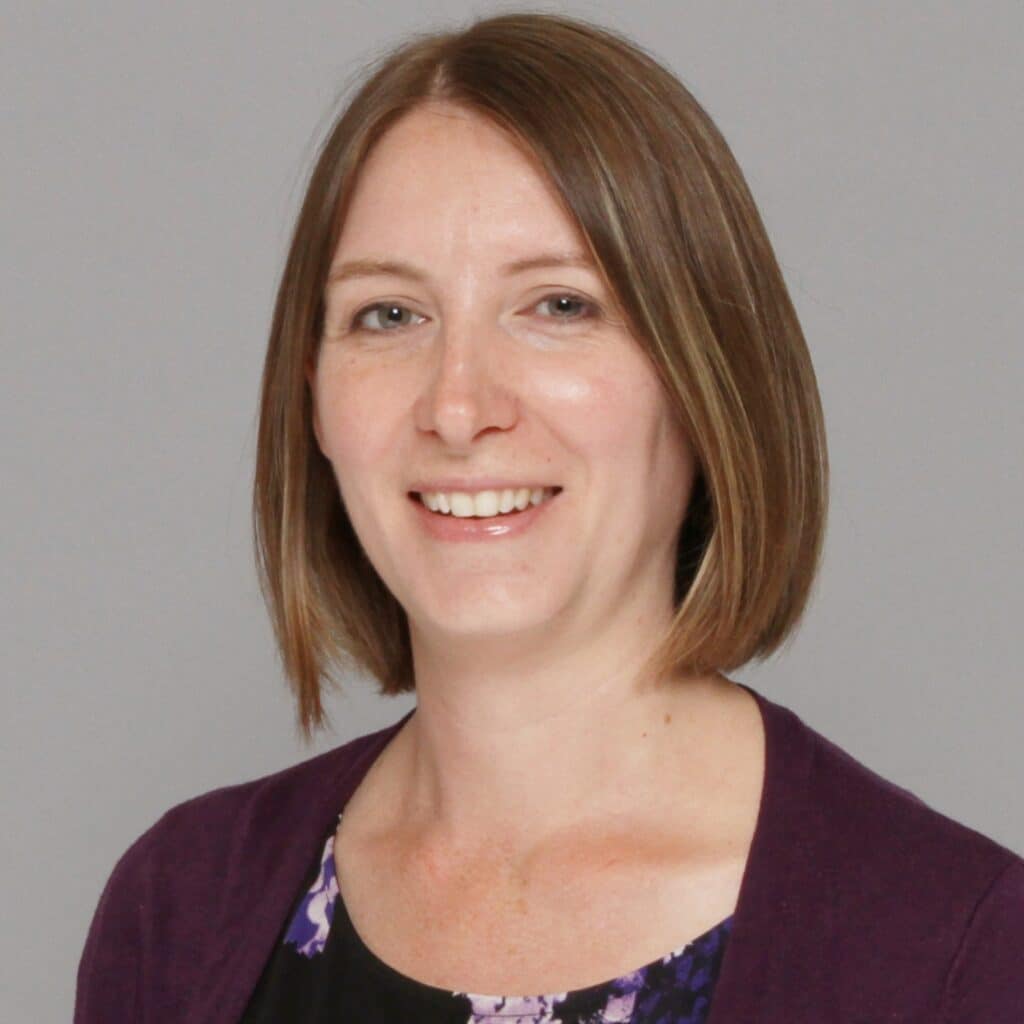 Kate Shuttleworth, a light-skinned female with light brown hair cut short to about the chin, wears a floral patterned shirt with a dark purple cardigan overtop, and smiles into the camera.