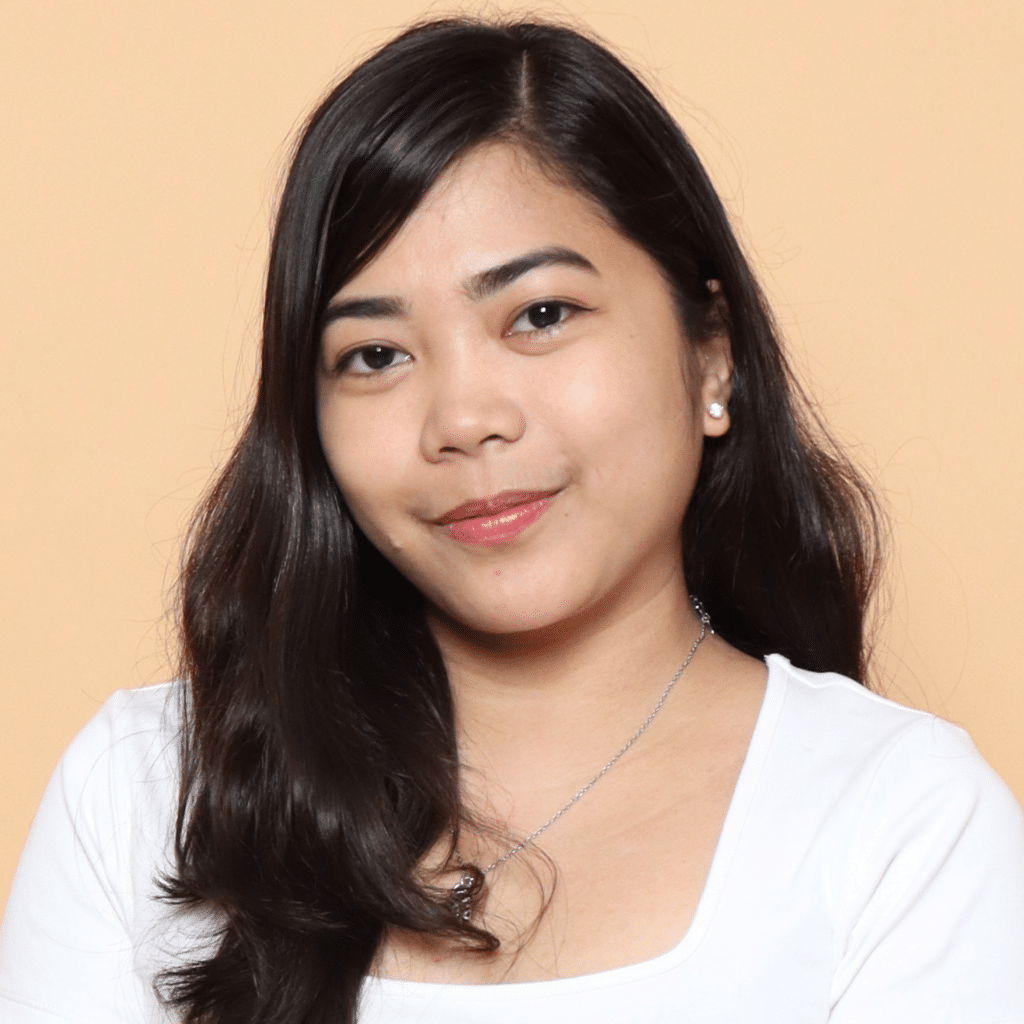 Blesilda's long, straight, dark hair cascades around her smiling face. Her dark brown eyes are framed with a tilted head, thoughtful expression, and smile reflecting curiosity. A white blouse-like t-shirt with a classic square neck cut frames her shoulders against a peach background.