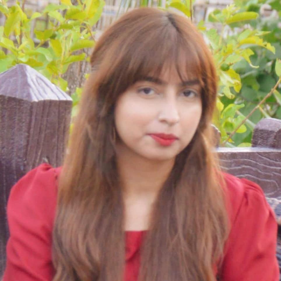 Hafsa Naeem is sitting outside under nice weather with green canopy in the background. Her long brown hair rests on her shoulders, which are adorned with a red dress.