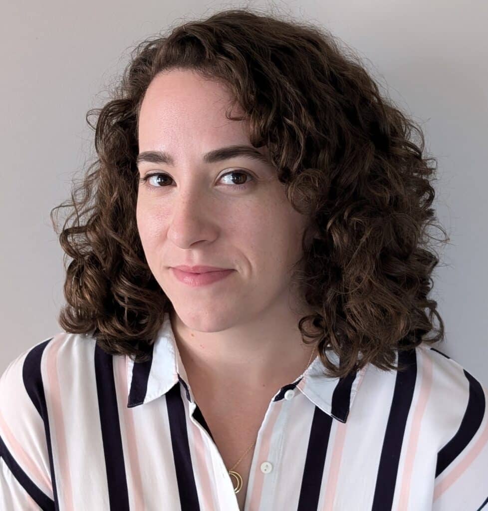 Kaitlin's dark brown, curly hair falls to her shoulders, on to a white blouse with a collar and black stripes with peach accents. She tilts her head to the side as she looks thoughtfully yet mysteriously into the camera, with a grin.