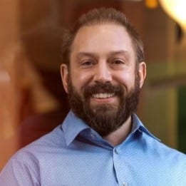 Beau poses in front of a window reflecting faded out random shapes. His short brown hair, moustache, and beard frame his smiling face, and his blue collar button-up shirt goes well with the brownish hues of the background.